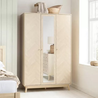 The CHOICEWARDROBE Helena 3 Door Mirrored Wardrobe features an oak chevron finish, wooden legs, and a full-length center mirror. Two baskets and a folded blanket rest on top, positioned by the window in a bright bedroom.