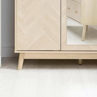 The CHOICEWARDROBE Helena 3 Door Mirrored Wardrobe features an oak chevron finish, angled wooden legs, and a full-length mirrored door that reflects drawers with matching wood grain and elegant legs.