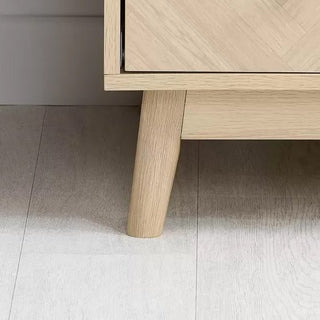 A close-up of the wooden leg and bottom corner of the CHOICEWARDROBE Helena 3 Door Mirrored Wardrobe in an oak chevron finish, set on a light-colored wooden floor.