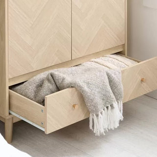 The CHOICEWARDROBE Helena 90cm 2 Door Wooden Wardrobe features a natural oak finish, chevron pattern front, and Scandinavian tapered legs, with its lower drawer open to show a folded beige and white fringed blanket inside.