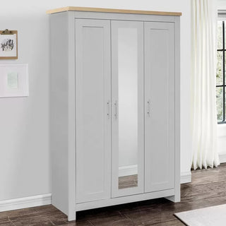 The CHOICEWARDROBE Highland 3 Door Wardrobe in grey with an oak top and mirrored center door (112cm wide) stands against a white wall near a window with white curtains.