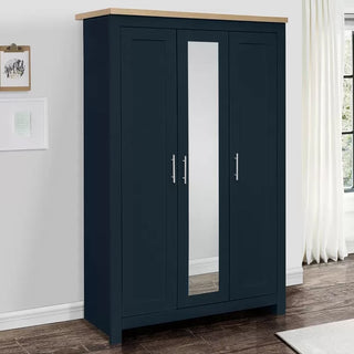 The CHOICEWARDROBE Highland 3 Door Wardrobe – Navy Blue with Oak Top & Mirror (112cm wide) stands against a white wall, near artwork and a window with white curtains, featuring a mirrored middle door and sleek silver handles.