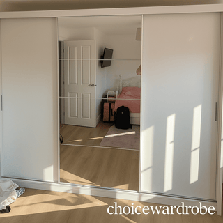 A CHOICEWARDROBE Inverto 3 Door Sliding Wardrobe (250cm, matt white, central mirror) stands in a wood-floored bedroom. The mirror reflects pink bedding on a bed, a door, wall-mounted TV, and personal items.