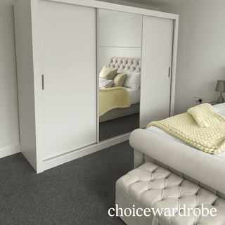 The CHOICEWARDROBE Inverto 3 Door Sliding Wardrobe, 250cm wide in matt white with a central mirror, stands in a bedroom with a beige upholstered bed, knitted yellow blanket, matching bench, and gray carpet.