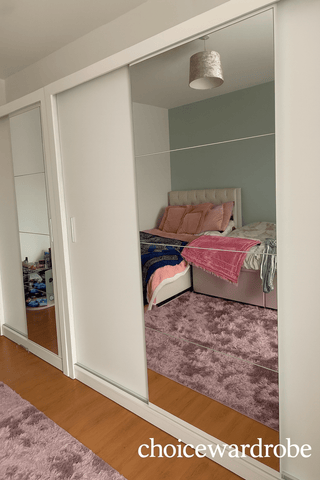 The CHOICEWARDROBE Inverto 3 Door Sliding Wardrobe (250cm wide, matt white with central mirror, built-in shelves & double hanging rail) reflects a neatly made bed with pink and blue bedding in a room with wooden flooring and pink carpet.