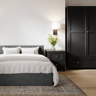 A modern bedroom features a neatly made bed with white bedding and a gray upholstered headboard. Next to the bed is a black nightstand with green flowers, and to the right stands a CHOICEWARDROBE Iowa 2 Door Wooden Wardrobe in matte black.