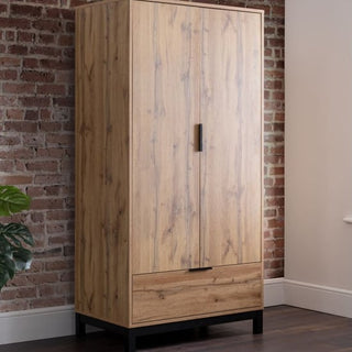 The CHOICEWARDROBE Kairo 2 Door 1 Drawer Wardrobe in rustic oak finish stands by a brick wall. With industrial style, black legs, two doors, a central handle, and bottom drawer, it's placed near a green plant for a cozy bedroom look.