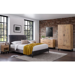 A modern bedroom with a gray upholstered bed, yellow throw blanket, wood nightstand, dresser, CHOICEWARDROBE Kairo 2 Door 1 Drawer Wardrobe in rustic oak finish, gray rug, brick accent wall, large windows, and framed wall art.