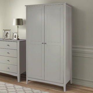 The CHOICEWARDROBE Kamuy Grey 2 Door Wardrobe in classic shaker style stands on a wooden floor beside a matching dresser, lamp, and framed photos. Light walls and simple molding complete the elegant scene.