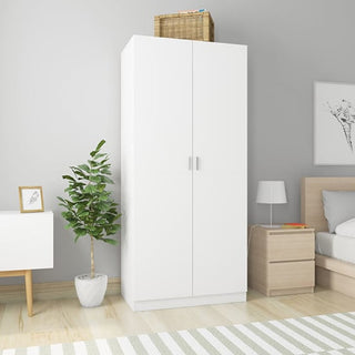 The CHOICEWARDROBE Kaylor White Wooden 2 Door Wardrobe adds sleek, minimal storage to a modern bedroom, placed beside a wooden nightstand and bed. A wicker basket sits on top, with a potted plant and white dresser nearby.