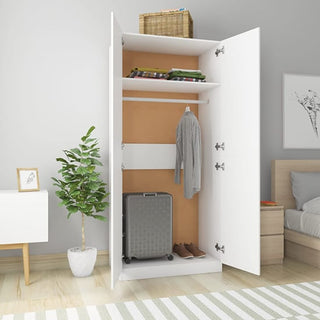 The CHOICEWARDROBE Kaylor White Wooden 2 Door Wardrobe stands open, displaying shelves with folded clothes, a wicker basket, a hanging shirt, and a gray suitcase at the bottom in a tidy bedroom with a plant and framed art.