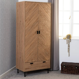 The CHOICEWARDROBE Lagos Wooden Wardrobe in oak features 2 doors, 1 drawer, a chevron pattern front, black metal legs, and a modern storage design. It stands next to grey curtains with a brown woven chest and beaded necklace nearby.