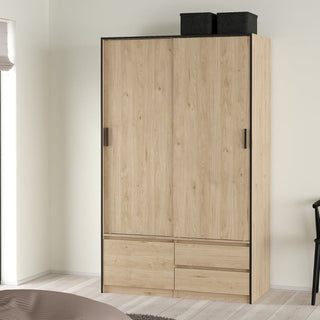 The CHOICEWARDROBE Lebanon Oak Sliding Door Wardrobe, featuring 2 doors and 2 drawers in an industrial modern style, stands against a cream bedroom wall, with part of a bed and chair nearby adding a cozy touch.