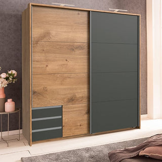 The CHOICEWARDROBE Limoni 2 Door Sliding Wardrobe in artisan oak and matt graphite offers contemporary bedroom storage with sliding doors and compact drawers. A nearby table with a flower vase completes the stylish look.