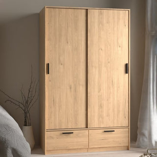 The CHOICEWARDROBE Liston 121cm Oak Finish Sliding Wardrobe with 2 doors, 2 drawers, and black handles stands in a bedroom by a window with sheer curtains and a vase holding tall branches.