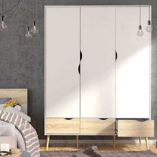 A modern bedroom features a light wood bed with gray bedding and the CHOICEWARDROBE Lorma 3 Piece Bedroom Furniture Set in white & oak, which includes a 3 door wardrobe, chest of drawers, and bedside table. Hanging bulbs complement the gray wall.