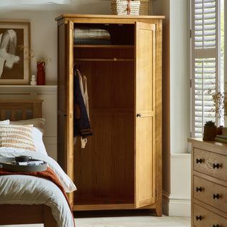 The CHOICEWARDROBE Mabli 97 cm 2 Door Waxed Oak Wardrobe with a solid wooden frame and classic Shaker design stands in a bright bedroom, open to show folded blankets and jackets alongside a bed, dresser, and window with white shutters.