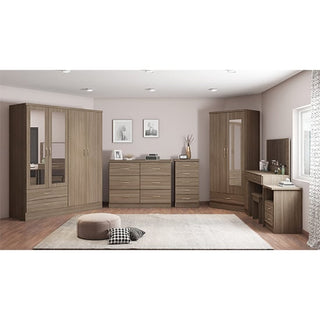 A modern bedroom with beige walls features the CHOICEWARDROBE Mack 4 Door Mirrored Wardrobe in Rustic Oak finish, providing spacious storage and full-length mirrors. A rug, pouf, and decor accents complete the tidy, well-lit space.