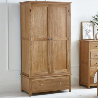 A CHOICEWARDROBE Macon 2 Door Solid Oak Wardrobe – Natural Finish, featuring drawer storage and a panelled design, stands against a white wall beside a matching chest of drawers with a vase and framed artwork on top.