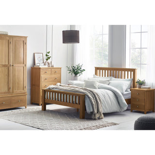 A bedroom features a wooden bed, white bedding, matching oak furniture, and the CHOICEWARDROBE Macon 3 Door Solid Oak Wardrobe with double drawer storage. A beige rug, large windows, green plants, and simple decor complete the charming look.