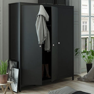 The CHOICEWARDROBE Macron 150 cm Black Wooden 3 Door Wardrobe stands in a bright room, its doors ajar. A gray hoodie hangs from one door, with plants and framed photos nearby, enhancing the modern bedroom storage setup.