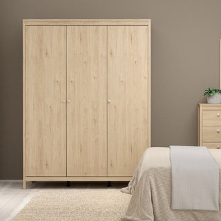 The CHOICEWARDROBE Macron 3 Door Wooden Wardrobe in an oak finish (150cm) with hanging rail and side shelves stands against a taupe wall, alongside a matching dresser, bed with beige bedding, and a plant.