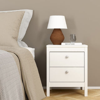 The CHOICEWARDROBE Macron 3 Piece Bedroom Furniture Set in white features a bedside table with two drawers next to a neatly made bed, topped with a lamp, glass of water, and the book SCULPTURE. Neutral tones complete the stylish look.