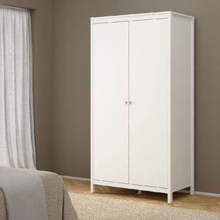 The CHOICEWARDROBE Macron 3 Piece Bedroom Furniture Set in white, featuring a 2 door wardrobe with simple metal handles, stands against a taupe wall. Part of a bed and a window with white curtains are seen nearby.