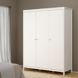 The CHOICEWARDROBE Macron 3 Piece Bedroom Set in classic white, featuring a 3 door wardrobe, stands against a beige wall with a white curtain on the left and a bed partially visible in the foreground.