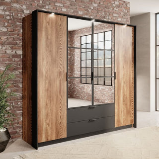 The CHOICEWARDROBE Madrid 223cm 4 Door Wooden Wardrobe in chestnut oak with a black frame, mirrors, drawers, and hanging rail stands against an exposed brick wall in a bright room with large windows.