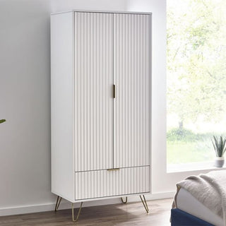 The CHOICEWARDROBE Marius Fluted 2 Door Wardrobe in a matt white finish with gold legs and drawer stands on a wooden floor beside a bed in a bright bedroom with a large window.