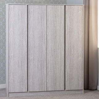 The CHOICEWARDROBE McGowan 150cm Wooden 4 Door Wardrobe in Light Grey Oak Finish stands on a carpeted floor against patterned wallpaper, beside a beige curtain, adding modern storage to the bedroom.
