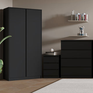 A room features the CHOICEWARDROBE McGowan 3 Piece Bedroom Set in black (2 door wardrobe, 4 drawer chest, bedside table). A white floating shelf holds books, a sculpture, a decorative figure, and a green plant.
