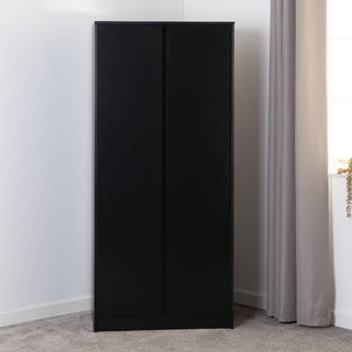 The CHOICEWARDROBE McGowen 4 Piece Black Wooden Bedroom Furniture Set, featuring a sleek, handleless 2-door wardrobe, stands against a white wall in a carpeted room with beige curtains partially covering the window.