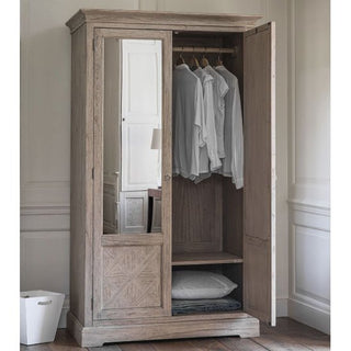 The CHOICEWARDROBE Mestiza 2 Door Wooden Wardrobe (111.5cm, natural finish) with a rustic mirrored design displays white shirts hanging and folded bedding inside, while a white laundry basket nearby adds style and practicality from Furniture in Fashion.