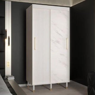 The CHOICEWARDROBE Millwall 100cm White Marble Effect Sliding Wardrobe, with gold handles and ribbed panels, stands in a modern bedroom. Its compact design features two sliding doors—one marble effect, one vertically grooved.