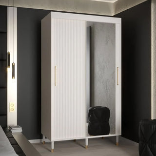 The CHOICEWARDROBE Millwall 120cm White Mirrored Sliding Door Wardrobe with fluted panels and gold handles stands against a dark wall, offering modern storage shelves. A black cushioned stool is placed in front of the stylish wardrobe.