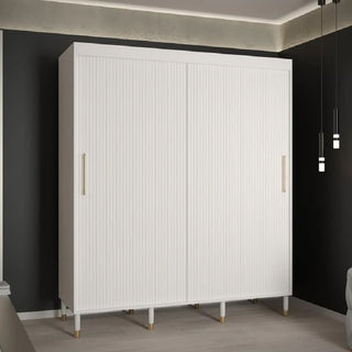 The CHOICEWARDROBE Millwall 180cm White Sliding 2 Door Wardrobe features ribbed panels and gold handles, standing against a dark wall in a modern room with light wooden flooring and elegant pendant lights for stylish bedroom storage.