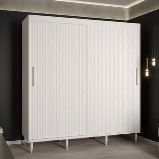 The CHOICEWARDROBE Millwall 200cm White Wooden Sliding Door Wardrobe features a fluted panel design, gold handles, and elevated legs, providing stylish and modern bedroom storage against a dark wall with pendant lighting.