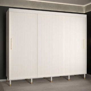 The CHOICEWARDROBE Millwall 250cm 3 Door Sliding Wardrobe in a white ribbed finish with gold handles stands against a dark wall on a light floor, illuminated by recessed lighting.