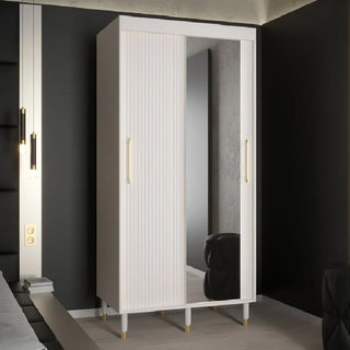 The CHOICEWARDROBE Millwall Mirrored Sliding Door Wardrobe, 100cm wide with a white finish, gold handles, and ribbed detailing, offers compact storage beside a bed in a modern bedroom with dark walls.