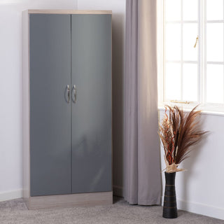 The CHOICEWARDROBE Modara Light Oak & Grey High Gloss 2 Door Wardrobe (78 cm) stands in a corner by a window with gray curtains; a black vase with dried plants sits on the floor beside it.