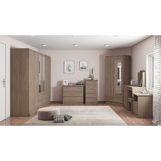 A bedroom with wood furniture, including the CHOICEWARDROBE Modara Rustic Oak Effect Mirrored Wardrobe (6 doors, 2 drawers), dressers, a vanity, light walls, wood floors, a rug, a pouf with plaid pillow, and two framed photos above the dresser.