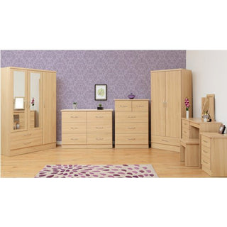 A bedroom with light wood furniture, including the CHOICEWARDROBE Modara Sonoma Oak Mirrored Wardrobe, chests, vanity table, and mirror. Purple patterned wallpaper and a leaf print rug add charm to the wooden floor.