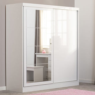 The CHOICEWARDROBE Modara White High Gloss Sliding Door Wardrobe with a mirrored front stands against a light gray wall, reflecting a white desk, stool, and pink decor. A pink rug adds a warm touch to the carpeted floor.