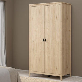 The CHOICEWARDROBE Nahlia Oak Wooden Wardrobe with two doors and black handles stands against a brown wall in a bedroom, alongside part of a bed, a window with white curtains, and a beige rug.