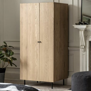 A CHOICEWARDROBE Nairo Natural Oak Wooden Wardrobe with 2 doors and black metal legs stands in a room with light walls, a plant, and part of a white fireplace nearby.