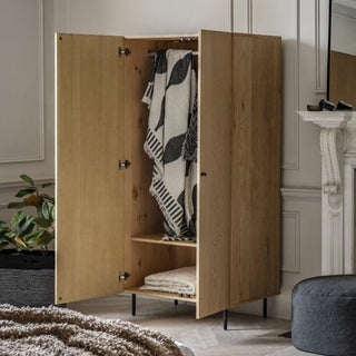 The CHOICEWARDROBE Nairo Natural Oak Wooden Wardrobe with 2 doors and black metal legs showcases hanging patterned blankets and a folded towel inside, blending modern Scandinavian style into a decoratively accented room with plants and furniture.