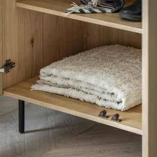 A folded textured cream blanket rests on the bottom shelf of a CHOICEWARDROBE Nairo Natural Oak Wooden Wardrobe with black metal legs. The 2-door wardrobe is open, and two small metal handles are on the shelf.