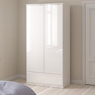 The CHOICEWARDROBE Nakou 3 Piece Bedroom Furniture Set in white high gloss features a modern 2 door wardrobe with lower drawer, set against wooden flooring and a light wall, with a striped blanket on the nearby bed.